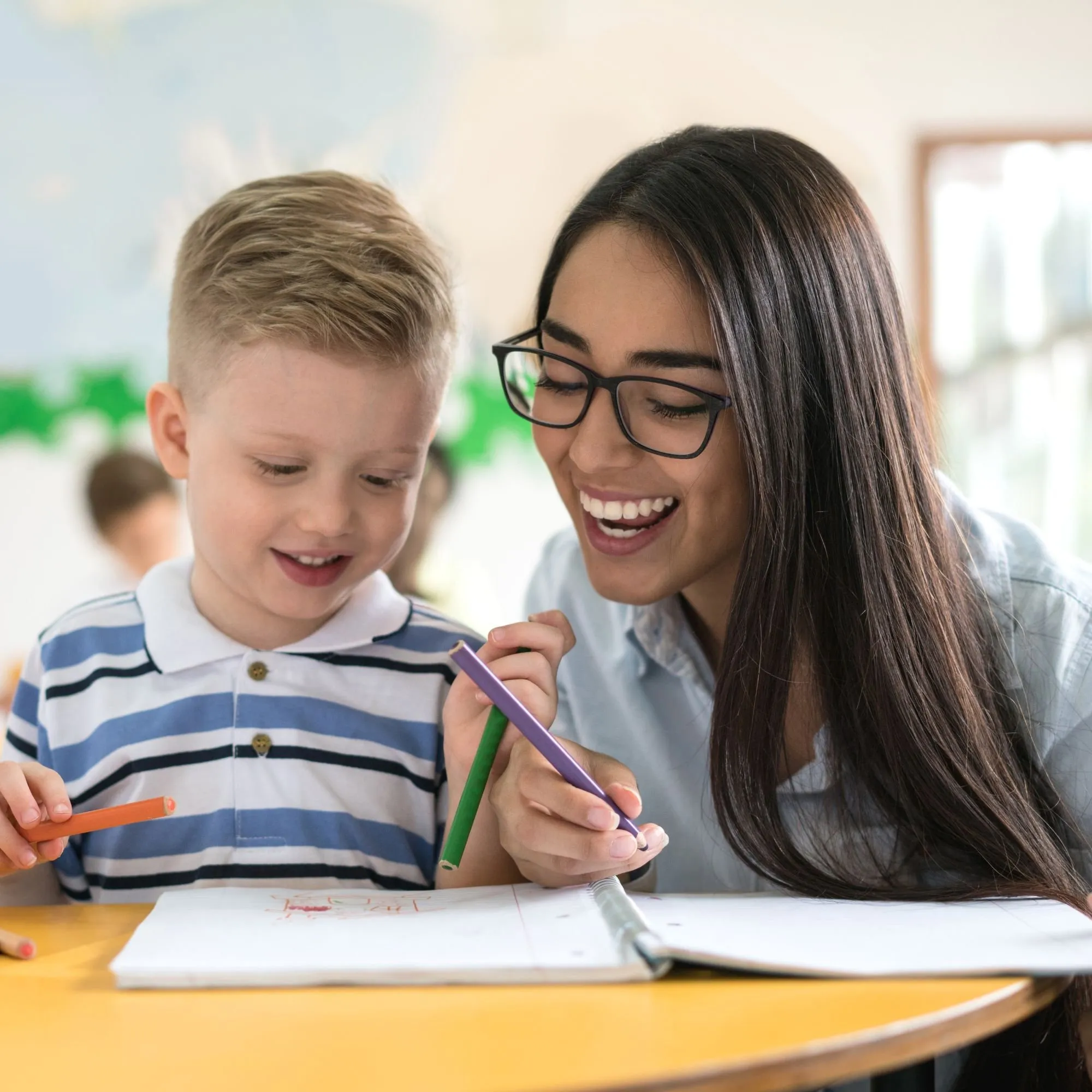 A teacher smiling as she helps a young student with his writing